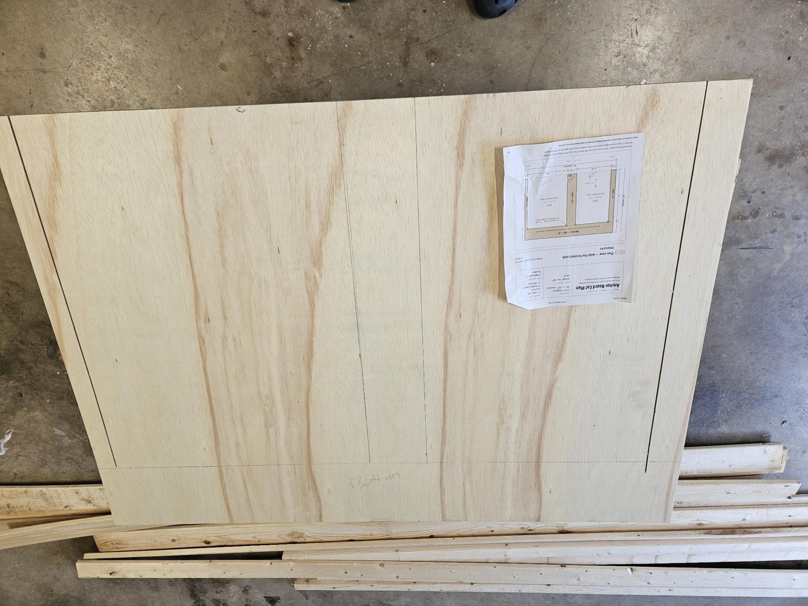 The anchor board blank on a garage floor with its strips and bridge line marked in pencil and the printed anchor board cut plan taped to it